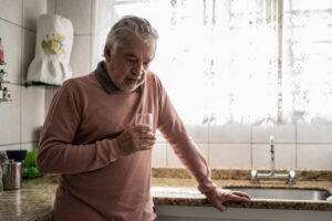 An older man stands in his kitchen drinking a glass of water, contemplating the connection between heart disease and depression.